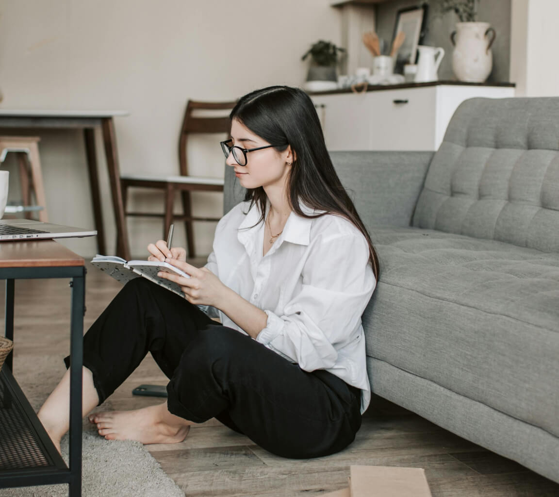 A woman writing in a journal leaning against a couch.